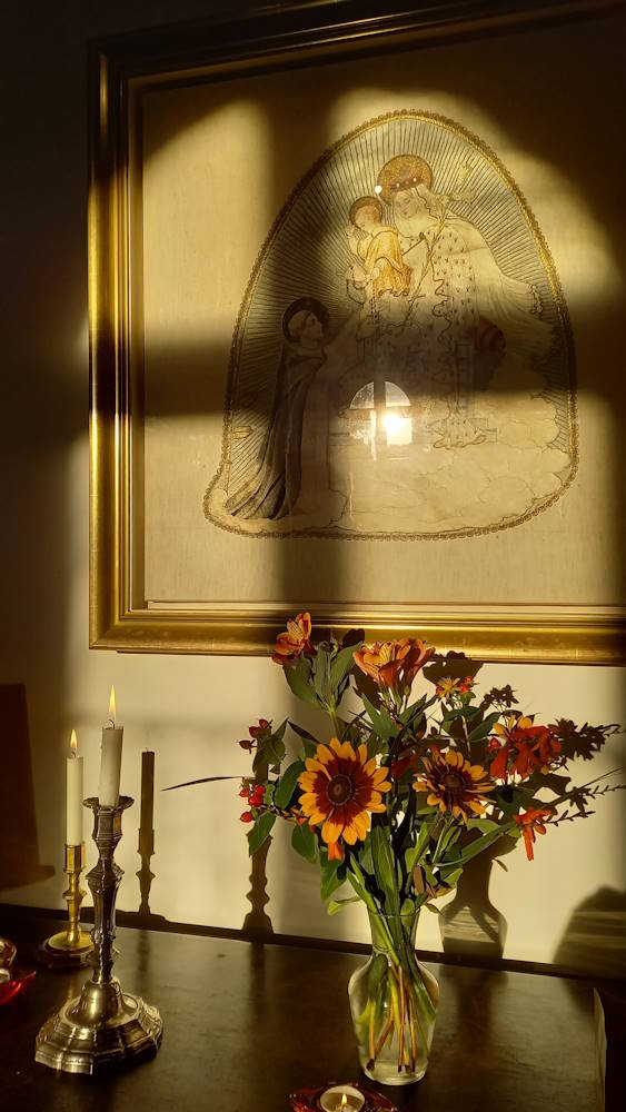 deux boigeoirs alumés et un vase avec un bouquet de fleurs sur un meuble collé au mur où se trouve un cadre représentant Saint Dominique au pied de la Vierge Marie avec l'Enfant Jésus dans ses bras